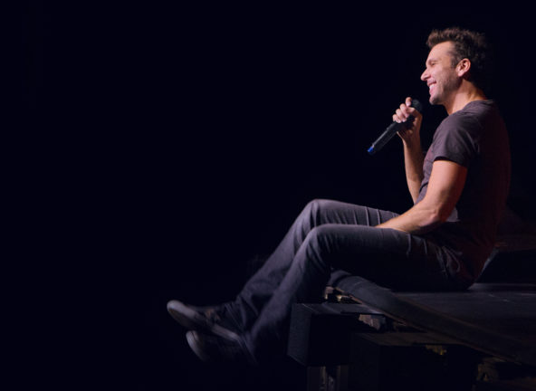 Dane Cook sitting on edge of stage grinning into a microphone