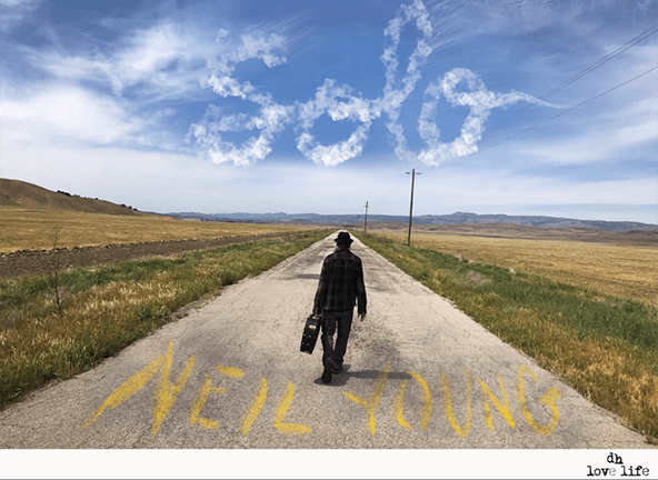 Neil Young walking solo with guitar down a straight north prairie road