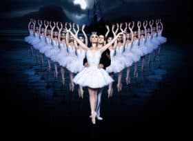 Ballet dancers in white dresses in v formation with lead dance couple, man and woman, at front