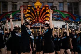 Students dressed in black tee shirts and masks doing a choreographed routine in front of a lit up colorful marquee of State Theatre.