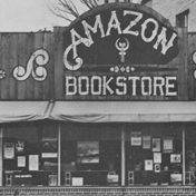 Storefront with large windows, awning over the sidewalk, and high arching sign above the one-story building facade with Amazon in arched lettering and Bookstore underneath with swirl designs on either side.