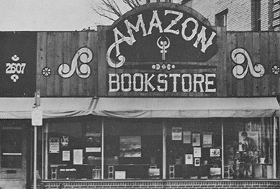 Storefront with large windows, awning over the sidewalk, and high arching sign above the one-story building facade with Amazon in arched lettering and Bookstore underneath with swirl designs on either side.
