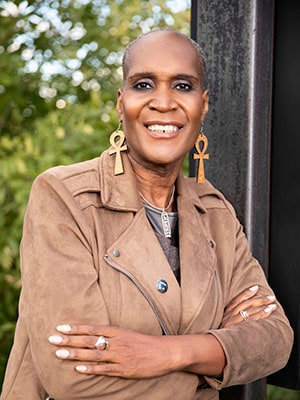 Andrea Jenkins in a light brown leather jacket with arms crossed in front of a large picture window.