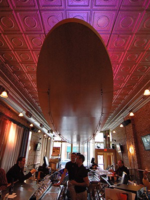 Boom! interior with view of bar, two bartenders in black shirts talking with a patron seated at the left, with large bright windows in the background at far end of room and colorful pink and purple light drenched tin ceiling with suspended oval wood fixture.