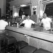 Two male bartenders in white short-sleeved shirts posing for a photo behind the bar with patrons seated on square leather stools, mostly congregated in far side and end of the bar.
