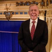 Fred Krohn in a black jacket and tartan red and green tie standing in the balcony of the Pantages Theatre with the stage and blue curtain in the background.