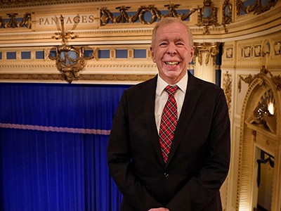 Fred Krohn in a black jacket and tartan red and green tie standing in the balcony of the Pantages Theatre with the stage and blue curtain in the background.