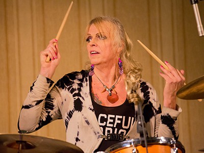 Jendeen Forberg wearing a black and white tie-died shirt playing a drumset in front of a beige theatre curtain.
