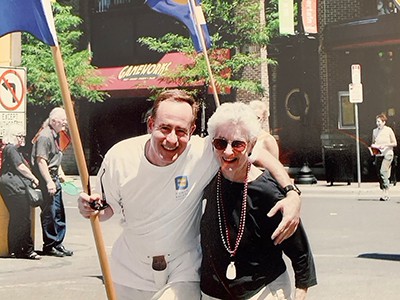 John Sullivan wearing white and flag pole belt with a flag and sunglasses in right hand and left arm around his mom, both leaning in smiling for the camera.