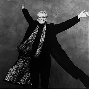 Lori Dokken wearing a flowing black jacket and leopard print shall with arms raised high and a large smile.