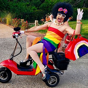 Miss Richfield wearing rainbow dress, platform heals, a black wig with colorful bowtie above white, thick-rimmed glasses seated on a red tricycle scooter on a gravel driveway.