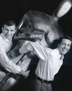 Scott Mayer and John Zeches on either side of a larger than life-sized Academy Award statue, tipped to the right as if they were carrying it off, both holding it from opposite sides.