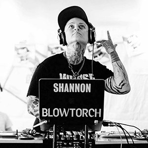 DJ Shannon Blowtorch with hat and large headphones on behind a turntable and compter with head tilted back and pointing upward with one hand.