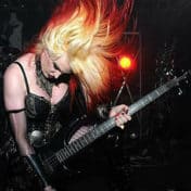 Tempest Crane wearing black lingerie playing a bass guitar and throwing cherry tipped, blonde hair skyward, caught in motion.