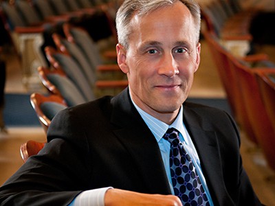 Tom Hoch in a black suit sitting in a theatre with arm over the back of the chair and rows of seats in the background.