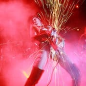 Venus De Mars on stage with smoke and red lighting and holding a microphone with sparks flying upward and downward.
