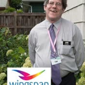 Tim G., wearing a gray shirt, a purple tie, a name tag on a lanyard, standing outside, with with the colorful bird design Wingspan logo superimposed over the left corner of the image.