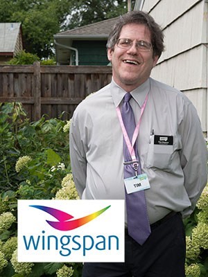 Tim G., wearing a gray shirt, a purple tie, a name tag on a lanyard, standing outside, with with the colorful bird design Wingspan logo superimposed over the left corner of the image.