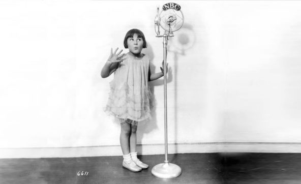 Girl in flowing dress poses with hands palms out next to NBC microphone that looms above her head. Photo Credit: Public Domain