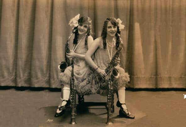 Conjoined sisters sit hip-to-hip on chair with saxophones in hand and two oboes standing on the ground in from them. Photo Credit: Hennepin County Public Library