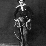 Man in chaps, black button-up shirt, white scarf around the neck, and hat, poses with a lasso in hand. Photo Credit Hennepin County Library