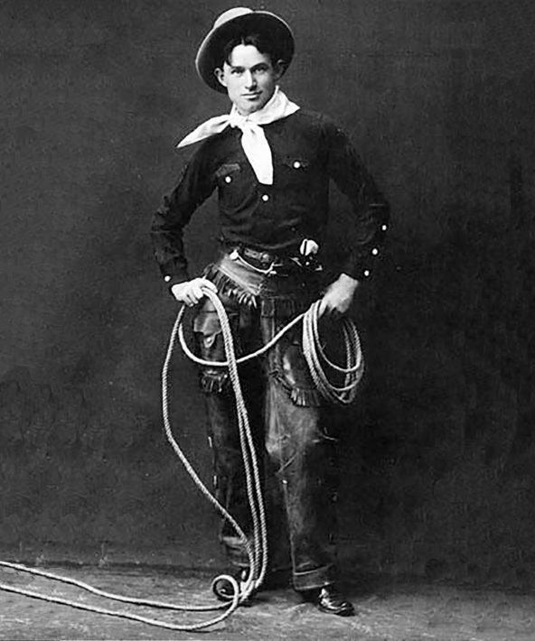Man in chaps, black button-up shirt, white scarf around the neck, and hat, poses with a lasso in hand. Photo Credit Hennepin County Library