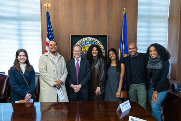 Hamilton cast, Spotlight students and Minnesota Secretary of State Steve Simon smiling and standing in front of the state seal, state flag, and American flag.