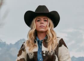 photo of Anne Wilson with blonde hair standing outside with mountains in the background. She's wearing a black cowgirl hat with a beige and brown cowprint jacket and blue denim shirt underneath.