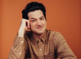 photo of Ben Schwartz wearing an orange button-down shirt, leaning on a table with his head leaning slightly against one of his hands.