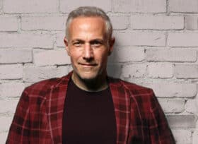 photo of Jim Brickman standing against a white brick wall wearing a red plaid jacket and black tshirt.