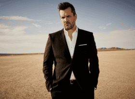 photo of Jim Jefferies standing in a remote desert wearing a dark suit and white open collar shirt with his hands in his jacket pockets.